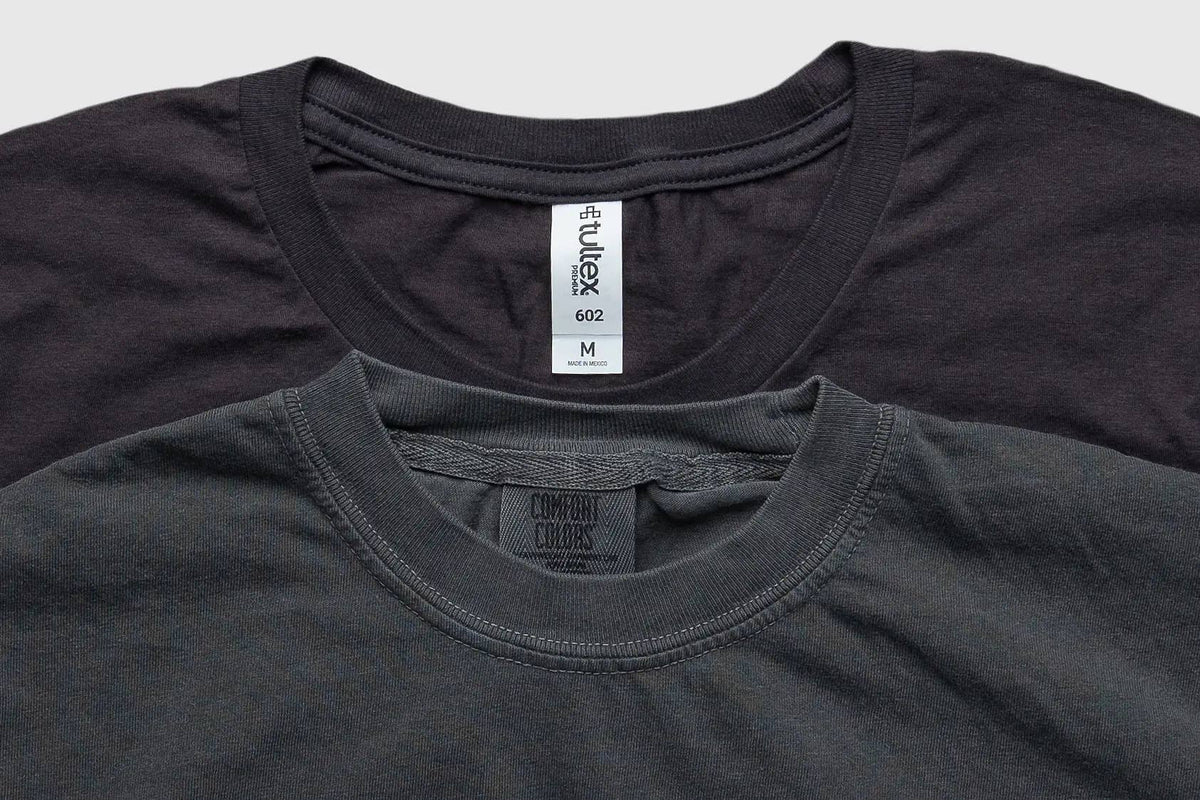 Two crewneck t-shirts stacked together, with a dark charcoal gray shirt in the foreground and a black shirt behind it. The foreground shirt shows a neck label for 'Comfort Colors' and the back shirt shows a neck label for 'Vulex'.