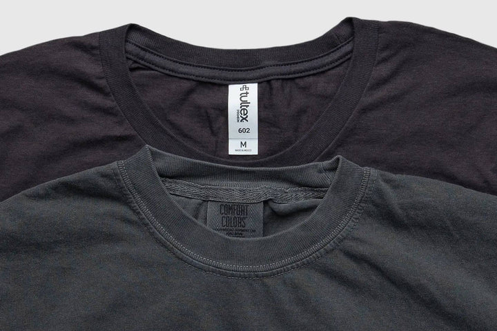 Two crewneck t-shirts stacked together, with a dark charcoal gray shirt in the foreground and a black shirt behind it. The foreground shirt shows a neck label for 'Comfort Colors' and the back shirt shows a neck label for 'Vulex'.