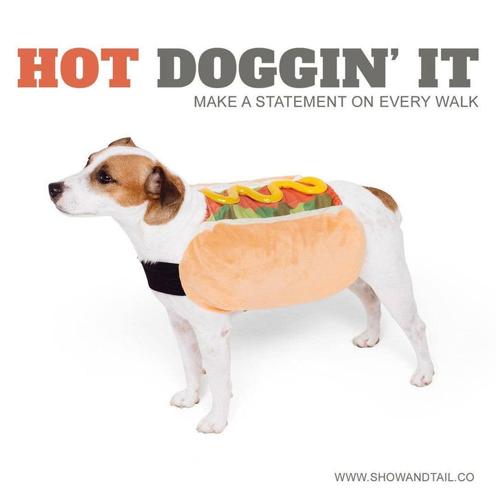 A small white and tan dog wearing a plush hot dog costume with a tan bun, green filling, yellow mustard, and a black belt, standing on a white background. Large text at the top reads 'HOT DOGGIN' IT' with smaller text 'MAKE A STATEMENT ON EVERY WALK', and 'WWW.