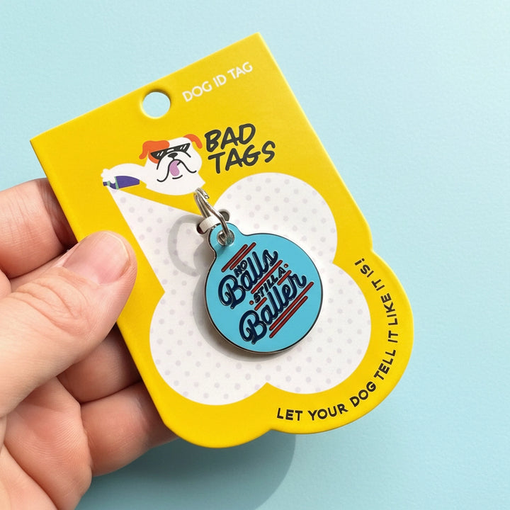 Dog ID tag with 'Balls to the Baller' text on a yellow card, held by a hand against a light blue background.