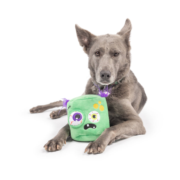 Gray dog resting with the Huxley & Kent PowerPlush Zombie dog toy positioned in front of its paws.